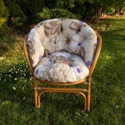 Poduszka na ratanowy fotel ogrodowy, kremowa z fioletowymi i brązowymi kwiatami, na trawie w ogrodzie.